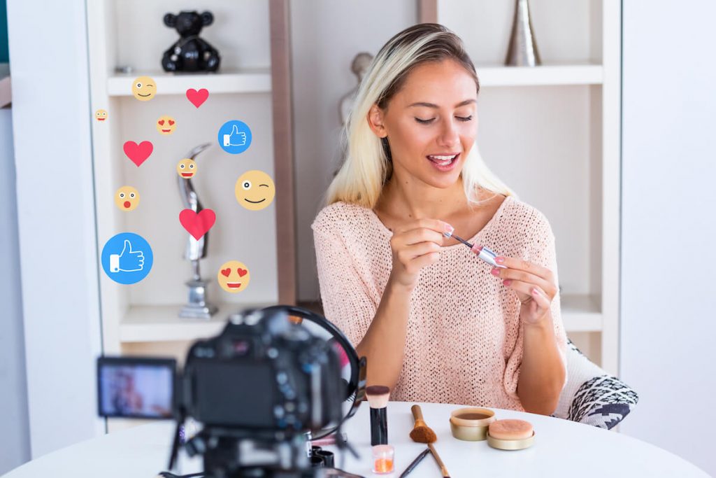 Benefits of influencer marketing: Woman putting on lipgloss with emojis floating above camera